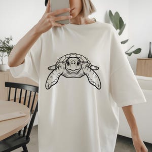May include: White oversized t-shirt featuring a black and white illustration of a sea turtle. The turtle design is centered on the front of the shirt. The shirt has short sleeves and a relaxed fit.