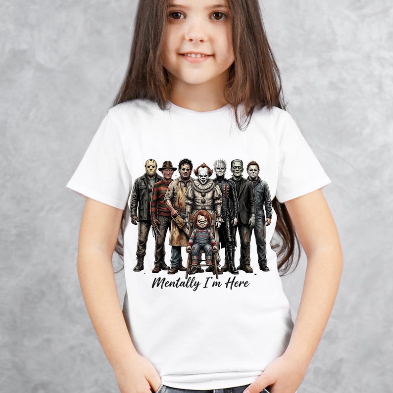 May include: A white short-sleeved t-shirt featuring a graphic print of various horror movie characters standing together, with a small doll figure sitting in front. The text "Mentally I'm Here" is printed in a black script font below the characters.