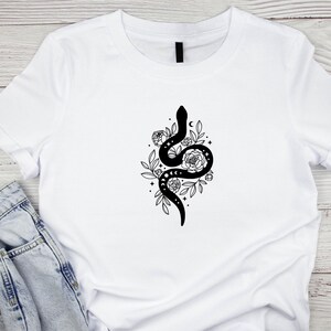 May include: White t-shirt featuring a black graphic of a snake intertwined with flowers, leaves, and moon phases. The design is centered on the shirt, creating a striking contrast against the white fabric. The shirt is made of a soft material.