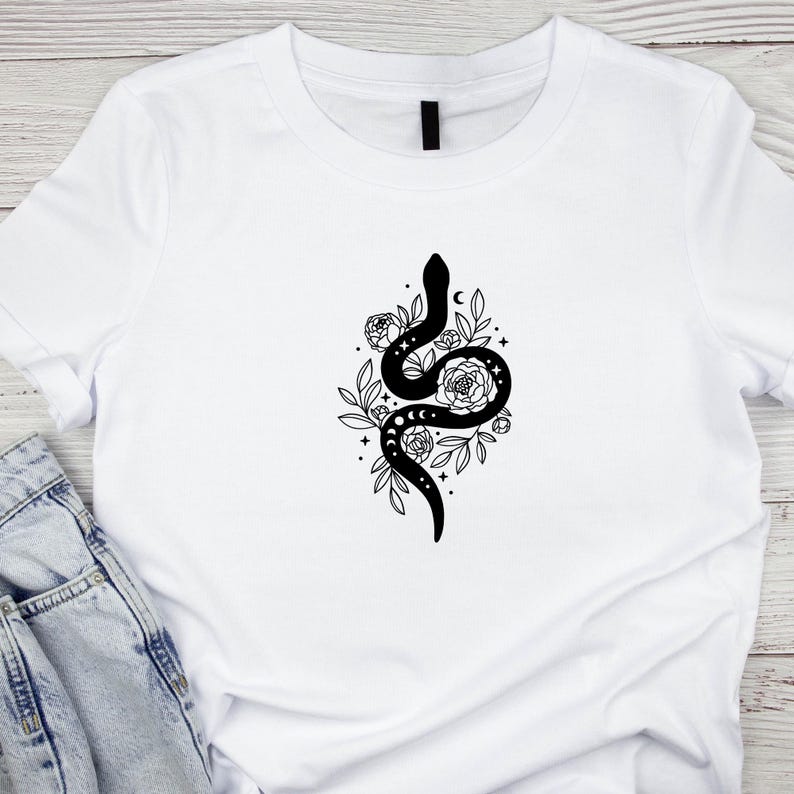 May include: White t-shirt featuring a black graphic of a snake intertwined with flowers, leaves, and moon phases. The design is centered on the shirt, creating a striking contrast against the white fabric. The shirt is made of a soft material.