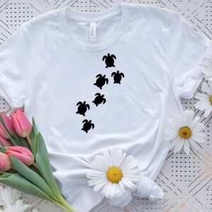 May include: White t-shirt featuring a design of six black sea turtles swimming diagonally across the front. The shirt is made of a soft, light-colored fabric. The design is simple and eye-catching.