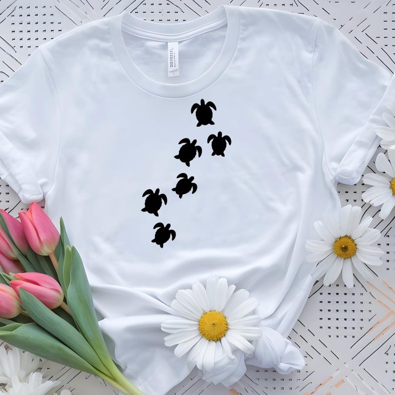 May include: White t-shirt featuring a design of six black sea turtles swimming diagonally across the front. The shirt is made of a soft, light-colored fabric. The design is simple and eye-catching.