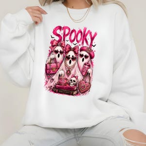 May include: A white long-sleeve sweatshirt featuring a graphic print of three ghosts wearing pink bows and heart-shaped sunglasses. The text SPOOKY appears above the ghosts, who are surrounded by pink accessories, books, and a skull.