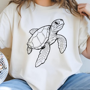 May include: Cream-colored t-shirt featuring a detailed black and white illustration of a sea turtle. The turtle is depicted with intricate shell patterns and flippers, giving a realistic appearance. The design is centered on the shirt.