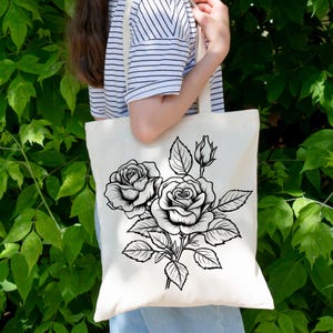 May include: Cream-colored tote bag featuring a black and white floral design of roses and leaves. The tote bag is made of a sturdy fabric and has a simple, elegant design. The bag is being held in front of a green leafy background.