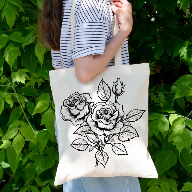 May include: Cream-colored tote bag featuring a black and white floral design of roses and leaves. The tote bag is made of a sturdy fabric and has a simple, elegant design. The bag is being held in front of a green leafy background.