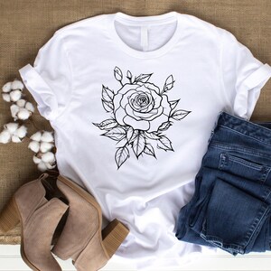 May include: White t-shirt featuring a black and white rose graphic. The rose design is surrounded by leaves. The shirt is paired with blue jeans and tan heeled boots. The image is taken on a neutral background.