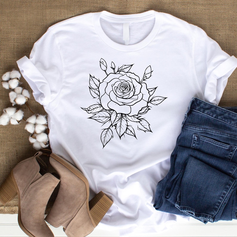 May include: White t-shirt featuring a black and white rose graphic. The rose design is surrounded by leaves. The shirt is paired with blue jeans and tan heeled boots. The image is taken on a neutral background.