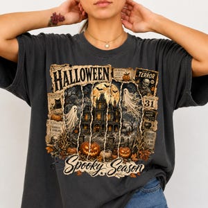 May include: A charcoal gray t-shirt featuring a graphic print with the text "HALLOWEEN" and "Spooky Season". The design includes a haunted house, ghosts, pumpkins, a black cat, a skeleton, and vintage-style newspaper clippings with "TALES OF TERROR".