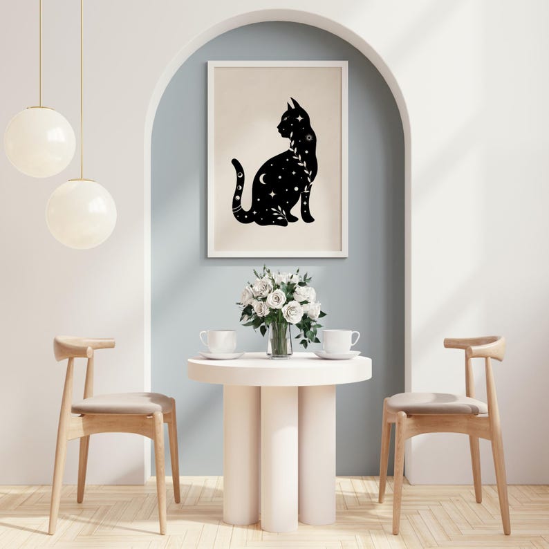 May include: A framed art print featuring a black cat silhouette adorned with celestial details like stars and a crescent moon. The artwork is displayed above a small table with a vase of white roses and two teacups. The room has a modern, minimalist design.