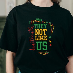 May include: Black t-shirt with a colorful graphic design. The design includes the words "Juneteenth" in red, "1865" in green, and the phrase "THEY NOT LIKE US" in green, yellow, and white. Stars and geometric shapes complete the design.