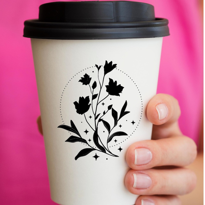 May include: A white paper coffee cup with a black lid. The cup features a black floral design inside a dotted circle, with leaves and star accents. The cup is held against a pink background.