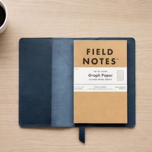 Può includere: Una copertina per notebook in pelle blu con un notebook di carta a quadretti Field Notes all'interno. Il notebook è aperto su una superficie di legno, con una tazza di caffè nell'angolo in alto a sinistra. Il notebook ha un segnalibro a nastro nero.