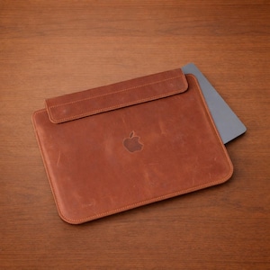 Può includere: Una custodia per laptop in pelle marrone con chiusura a patta e bordi cuciti. Il logo Apple è impresso sul davanti. Un laptop grigio è parzialmente visibile all'interno, su una superficie di legno.