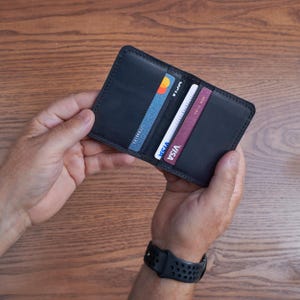 Op de afbeelding: Een zwart leren portemonnee, open om verschillende creditcards te tonen. De portemonnee wordt in twee handen gehouden, waarbij de binnenvakken en de kleurrijke ontwerpen van de kaarten worden getoond. De portemonnee lijkt nieuw en ongebruikt.