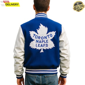 Op de afbeelding: Blauw en witte varsity jas met de woorden "TORONTO MAPLE LEAFS" in wit op de rug. De jas heeft witte mouwen en een wit esdoornbladlogo. De jas heeft een gestreepte kraag en manchetten.