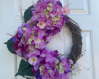 May include: A purple and green floral wreath with a grapevine base. The wreath is decorated with purple hydrangeas and pink flowers.