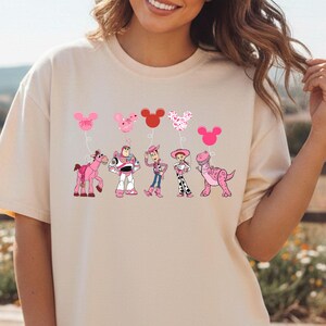 May include: Cream-colored t-shirt featuring a graphic of cartoon characters and heart-shaped balloons in pink and red. The design includes a pink horse, Buzz Lightyear, Woody, Jessie, and a dinosaur. The shirt is a relaxed fit.
