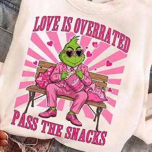 May include: White sweatshirt with a graphic of a green character wearing heart-shaped sunglasses and a pink suit, holding popcorn. The text "LOVE IS OVERRATED" and "PASS THE SNACKS" are printed in pink.