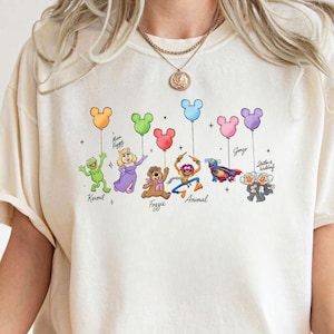 May include: Cream-colored t-shirt featuring a colorful graphic of Muppet characters holding balloons. The characters include Kermit, Miss Piggy, Fozzie, Animal, and others. The balloons are in various colors, and the names of the characters are written below them.