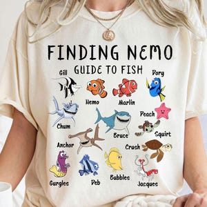 Puede incluir: Camiseta color crema con el texto "FINDING NEMO" y una guía de personajes de peces de la película. La guía incluye ilustraciones de Nemo, Dory, Marlin y otros personajes. La camiseta es de corte holgado.