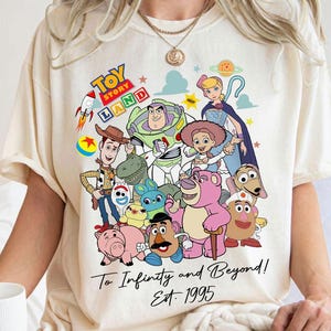 Puede incluir: Camiseta de color crema con un gráfico colorido de personajes de Toy Story, incluyendo a Woody, Buzz Lightyear y otros. La camiseta tiene el texto "Toy Story Land" y "To Infinity and Beyond! Est. 1995".