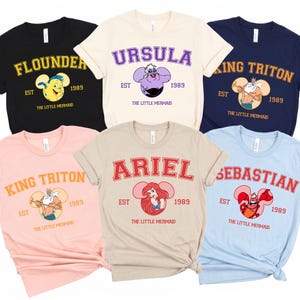 Può includere: Una collezione di magliette con personaggi de "La Sirenetta". Ogni maglietta mostra il nome di un personaggio in un font universitario, insieme a un'immagine del personaggio e l'anno 1989. Le magliette sono disponibili in vari colori, tra cui nero, crema, blu navy, rosa, beige e azzurro.