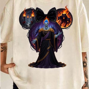 May include: Off-white t-shirt featuring a graphic of Hades from Disney's Hercules. The design includes a black bow, flames, and a three-headed dog. Hades is depicted with blue skin and a black robe.