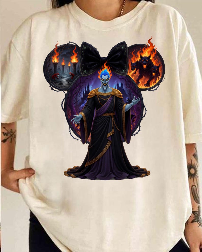 May include: Off-white t-shirt featuring a graphic of Hades from Disney's Hercules. The design includes a black bow, flames, and a three-headed dog. Hades is depicted with blue skin and a black robe.