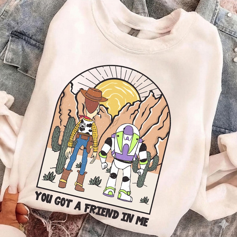 May include: White crewneck sweatshirt featuring a graphic of Woody and Buzz Lightyear in a desert scene with a sun and mountains. The text "YOU GOT A FRIEND IN ME" is printed at the bottom. The sweatshirt is made of a soft material.