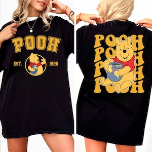 Pode incluir: T-shirt preto com um design amarelo e dourado. A frente exibe "POOH" e um urso de desenho animado com um pote de mel, com "EST. 1926". A parte de trás repete "POOH" com o urso. Uma parte de cima divertida e casual.