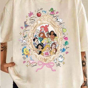 May include: Cream-colored t-shirt featuring a gold-framed oval design with various Disney princesses. The design includes illustrations of characters like Ariel, Cinderella, and Belle, along with their names. A pink bow is at the bottom.