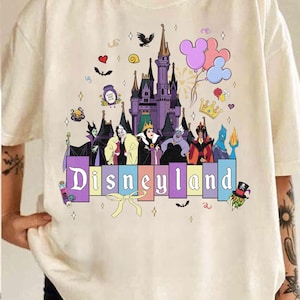 Pu&ograve; includere: T-shirt color crema con grafica Disneyland. Il design include un castello viola, cattivi Disney, palloncini di Topolino e la parola "Disneyland" in lettere colorate. Ulteriori elementi decorativi includono cuori, stelle e un fiocco.