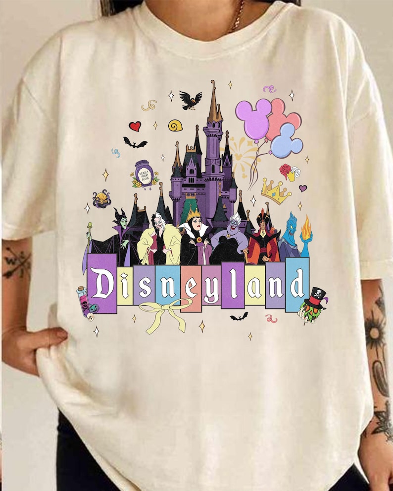 Pu&ograve; includere: T-shirt color crema con grafica Disneyland. Il design include un castello viola, cattivi Disney, palloncini di Topolino e la parola "Disneyland" in lettere colorate. Ulteriori elementi decorativi includono cuori, stelle e un fiocco.