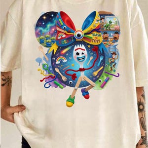 May include: Cream-colored t-shirt featuring a colorful graphic of the character Forky from the movie Toy Story 4. The design includes a bow, the words "Trash to Toy!" and other characters from the movie.