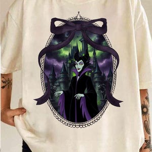 May include: Cream-colored t-shirt featuring a framed graphic of a character in a black and purple outfit with a castle backdrop. The graphic is framed by a black bow and an oval border. The character has green skin and black horns.
