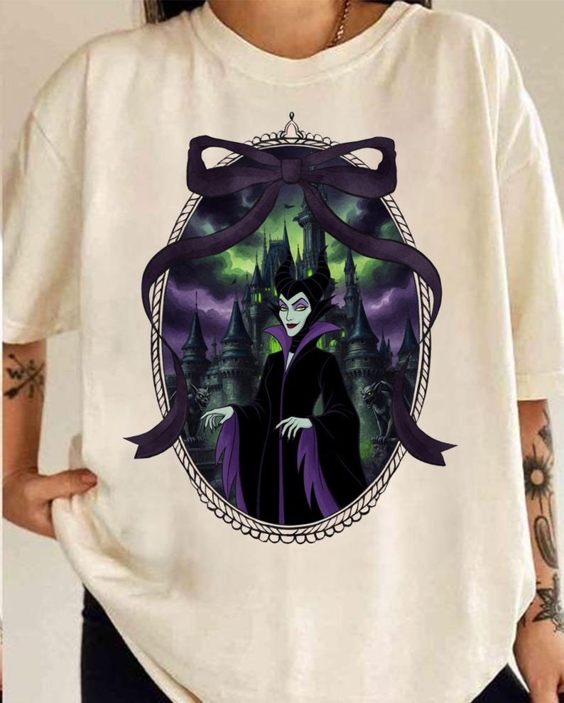 May include: Cream-colored t-shirt featuring a framed graphic of a character in a black and purple outfit with a castle backdrop. The graphic is framed by a black bow and an oval border. The character has green skin and black horns.