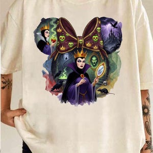 Puede incluir: Camiseta color crema con un gr&aacute;fico de un lazo con calaveras y coronas, e im&aacute;genes de una reina sosteniendo un espejo y una manzana. El dise&ntilde;o incluye un castillo, cuervos y una rata. La obra de arte es de estilo caricatura.