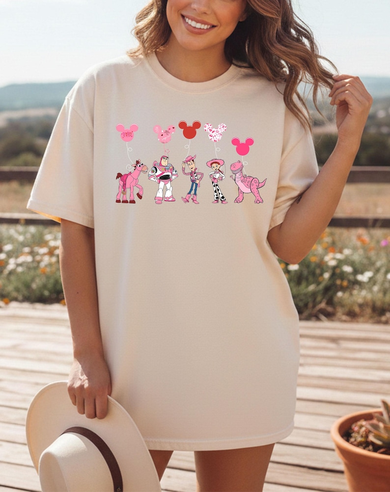 May include: Cream-colored t-shirt featuring a graphic of cartoon characters and heart-shaped balloons in pink and red. The design includes a pink horse, Buzz Lightyear, Woody, Jessie, and a dinosaur. The shirt is a relaxed fit.