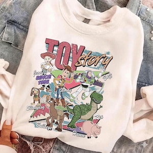 Puede incluir: Sudadera blanca de cuello redondo con un gr&aacute;fico colorido de los personajes de Toy Story, incluyendo a Woody, Buzz Lightyear y Rex. El dise&ntilde;o incluye el texto "Toy Story" y "Since 1995". La sudadera es de material suave.