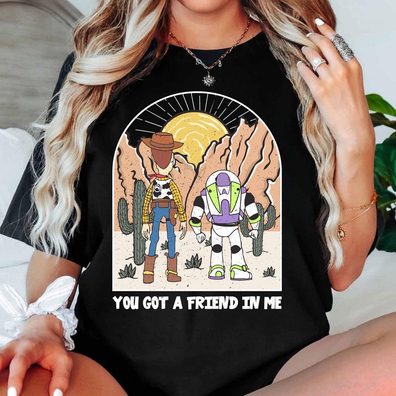 May include: Black t-shirt featuring a graphic of Woody and Buzz Lightyear in a desert scene with cacti and a sun. The text "YOU GOT A FRIEND IN ME" is printed below the characters. The design is in a cartoon style.