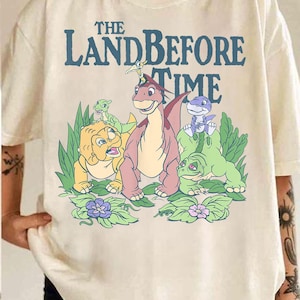 May include: Cream-colored t-shirt featuring the text "THE LAND BEFORE TIME" in blue above a graphic of cartoon dinosaurs. The graphic includes a pink, yellow, and green dinosaur with purple flowers and green leaves.