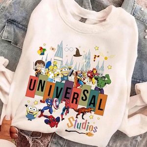 May include: White crewneck sweatshirt featuring a colorful graphic of various cartoon characters and the text "UNIVERSAL Studios". The design includes characters from Harry Potter, The Simpsons, Marvel, and more. The sweatshirt is on a denim background.