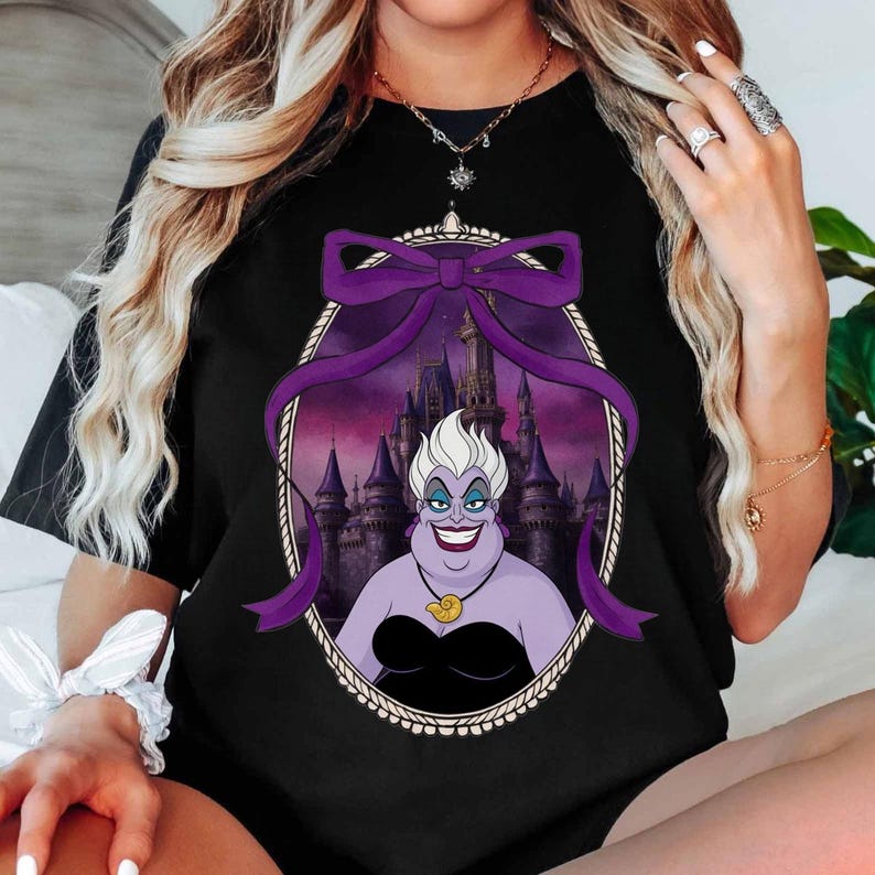 May include: Black t-shirt featuring a cartoon illustration of Ursula from The Little Mermaid. The image shows Ursula with a purple bow and a castle backdrop. The design is framed in an oval with a decorative border.