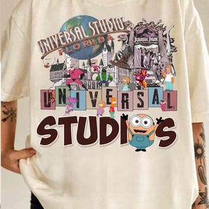 May include: Cream-colored t-shirt featuring a colorful graphic of Universal Studios Florida. The design includes the park's logo, various cartoon characters, and the words "UNIVERSAL STUDIOS FLORIDA" and "STUDIOS". A Minion character is also featured.