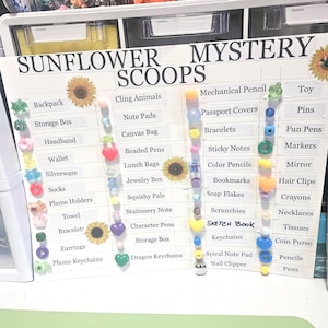 May include: A white board with the words "SUNFLOWER MYSTERY SCOOPS" and a list of items like backpacks, bracelets, and keychains. The board has decorative sunflowers and colorful beads attached to it.