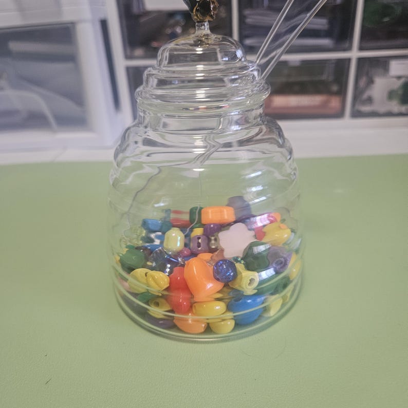 May include: A clear glass jar with a lid and a spoon, filled with colorful beads. The beads are various shapes and colors, including yellow, orange, blue, and green. The jar is sitting on a light green surface.