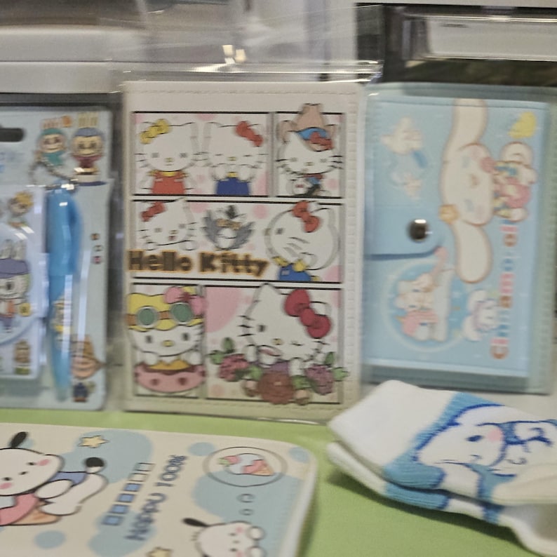 May include: A collection of Hello Kitty and Sanrio-themed stationery and accessories. Includes a Hello Kitty comic-style notebook, a light blue pen, a light blue notebook with a button closure, and a pair of white socks with a blue character design.