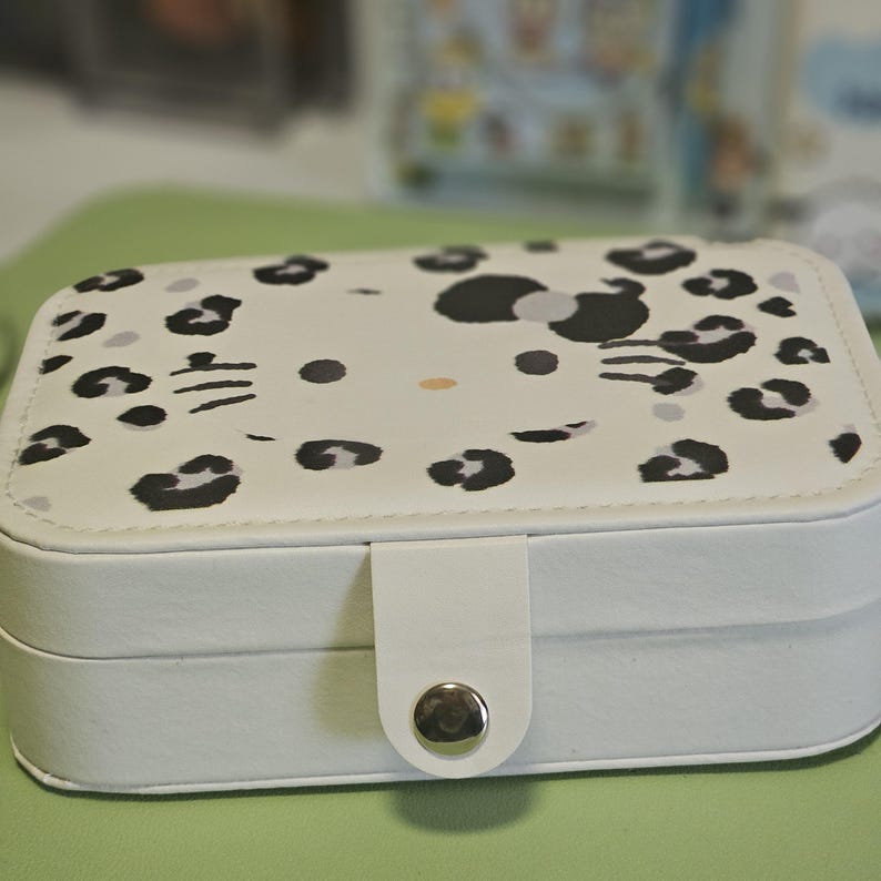 May include: A white rectangular jewelry box with a lid featuring a pattern of black leopard spots and a cartoon cat face. The box has a white clasp with a silver button closure. The box is on a green surface.