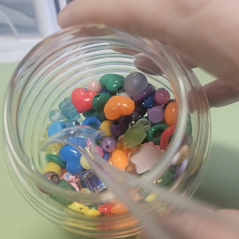 May include: A clear glass jar filled with colorful beads of various shapes and sizes, including hearts, stars, and round beads. The beads are in shades of red, orange, green, blue, purple, and pink.